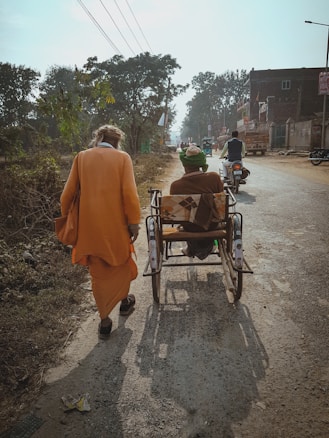 An older adult dressed in orange walks beside a person in a wheelchair on a rural road. The road is lined with trees and buildings. Other people are seen riding motorcycles further ahead.