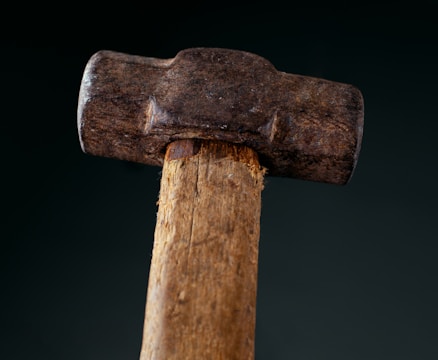 A close-up view of a worn hammer with a wooden handle and a metal head. The metal head shows signs of rust and wear, indicating frequent use. The wooden handle is slightly splintered and rough to the touch.