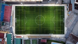 An aerial view of a rectangular soccer field situated in an urban area. The field is outlined with white and yellow lines, showing a well-defined layout for a soccer game. The green artificial turf is surrounded by various buildings and structures on all sides, giving the atmosphere of a compact urban setting. The floodlights indicate potential for evening use.