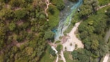 An aerial view of a lush green landscape with dense forests surrounding a river or stream. The water appears clear with blue and green tones, and there are visible pathways and sandy areas nearby. A small structure is situated near the water.