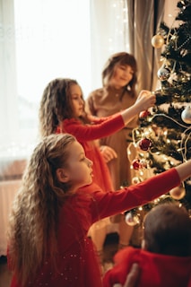 Children are decorating a Christmas tree with ornaments and lights in a warmly lit room. The tree has various decorations, including shiny baubles. The children are dressed in festive clothing, with one having a red dress.