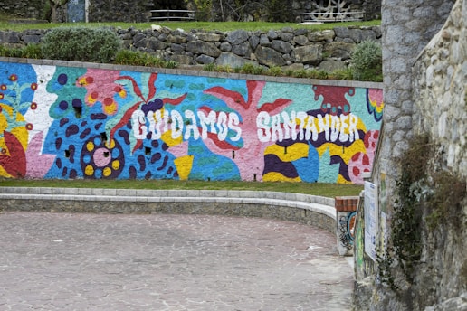 A vibrantly painted mural on a stone wall features abstract shapes and patterns in vivid colors. The text 'CUIDAMOS SANTANDER' is prominently displayed in white against a lively backdrop that incorporates a mix of blue, pink, green, yellow, and red. The setting includes cobblestone pavement in the foreground and stone walls with greenery visible above the mural.