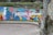A vibrantly painted mural on a stone wall features abstract shapes and patterns in vivid colors. The text 'CUIDAMOS SANTANDER' is prominently displayed in white against a lively backdrop that incorporates a mix of blue, pink, green, yellow, and red. The setting includes cobblestone pavement in the foreground and stone walls with greenery visible above the mural.