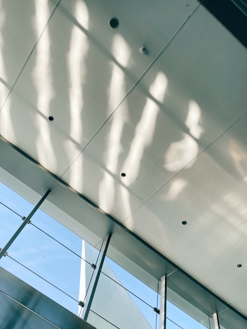 Light streams through a large, modern glass facade, casting soft, wavy shadows on the ceiling. The architecture is characterized by sleek lines and open spaces, and the contrast between the blue sky and the interior adds a serene atmosphere.
