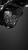 A glossy black liquid appears to be in motion, capturing the fluid dynamics of a droplet about to detach from a larger mass. The surface is reflective, and the liquid forms intricate patterns and waves, conveying a sense of movement and fluidity.