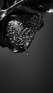 A glossy black liquid appears to be in motion, capturing the fluid dynamics of a droplet about to detach from a larger mass. The surface is reflective, and the liquid forms intricate patterns and waves, conveying a sense of movement and fluidity.