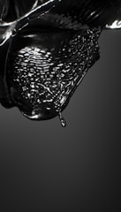 A glossy black liquid appears to be in motion, capturing the fluid dynamics of a droplet about to detach from a larger mass. The surface is reflective, and the liquid forms intricate patterns and waves, conveying a sense of movement and fluidity.