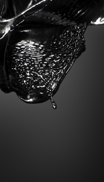 A glossy black liquid appears to be in motion, capturing the fluid dynamics of a droplet about to detach from a larger mass. The surface is reflective, and the liquid forms intricate patterns and waves, conveying a sense of movement and fluidity.