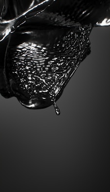A glossy black liquid appears to be in motion, capturing the fluid dynamics of a droplet about to detach from a larger mass. The surface is reflective, and the liquid forms intricate patterns and waves, conveying a sense of movement and fluidity.