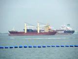 Two large cargo ships are navigating through the water. The ship closer to the viewer has a prominent red hull and yellow cranes, while the ship in the background has the word 'TANKER' visible on its side. The sea is calm with a series of blue buoys in the foreground.