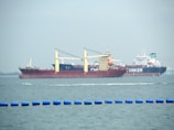 Two large cargo ships are navigating through the water. The ship closer to the viewer has a prominent red hull and yellow cranes, while the ship in the background has the word 'TANKER' visible on its side. The sea is calm with a series of blue buoys in the foreground.
