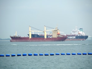 Two large cargo ships are navigating through the water. The ship closer to the viewer has a prominent red hull and yellow cranes, while the ship in the background has the word 'TANKER' visible on its side. The sea is calm with a series of blue buoys in the foreground.