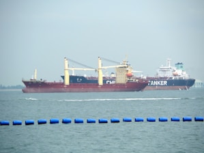Two large cargo ships are navigating through the water. The ship closer to the viewer has a prominent red hull and yellow cranes, while the ship in the background has the word 'TANKER' visible on its side. The sea is calm with a series of blue buoys in the foreground.