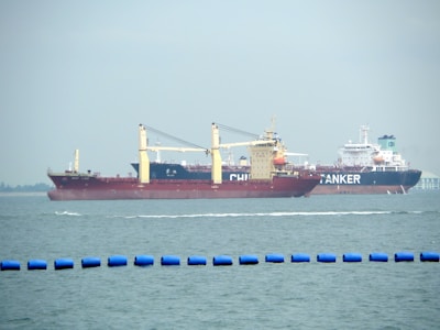 Two large cargo ships are navigating through the water. The ship closer to the viewer has a prominent red hull and yellow cranes, while the ship in the background has the word 'TANKER' visible on its side. The sea is calm with a series of blue buoys in the foreground.