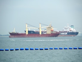 Two large cargo ships are navigating through the water. The ship closer to the viewer has a prominent red hull and yellow cranes, while the ship in the background has the word 'TANKER' visible on its side. The sea is calm with a series of blue buoys in the foreground.