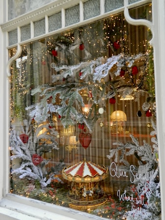 A festive window display featuring snow-covered pine branches, red ornaments, and a miniature carousel figurine surrounded by twinkling lights. A handwritten phrase 'Que Sera Sera, What will be will be' is visible on the glass. Warm lights and reflections from inside create a cozy ambiance.