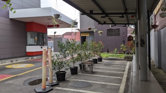 A small outdoor space with potted plants arranged neatly along the edge of a sheltered area. The pavement is marked with parking lines, and a security booth with a large overhang is visible to the left. Above the area, there are additional plants hanging. The surroundings include a building with various architectural features and a sign that reads 'Musholla'.