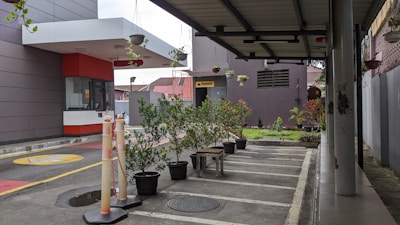 A small outdoor space with potted plants arranged neatly along the edge of a sheltered area. The pavement is marked with parking lines, and a security booth with a large overhang is visible to the left. Above the area, there are additional plants hanging. The surroundings include a building with various architectural features and a sign that reads 'Musholla'.