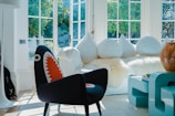 A brightly lit living room features a modern interior design with a prominent plush white couch adorned with round, white cushions. A unique black chair with a colorful shark-mouth design stands out in the foreground. Large windows fill the space with natural light, illuminating the room. A light blue, modern coffee table is visible alongside a decorative, large rattan vase.