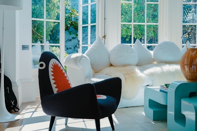 A brightly lit living room features a modern interior design with a prominent plush white couch adorned with round, white cushions. A unique black chair with a colorful shark-mouth design stands out in the foreground. Large windows fill the space with natural light, illuminating the room. A light blue, modern coffee table is visible alongside a decorative, large rattan vase.