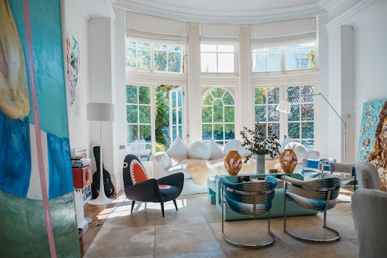 A bright and airy living room with large, arched windows letting in abundant natural light. The space is stylishly decorated with modern furniture, including a sofa with white cushions, unique armchairs, and a quirky shark-themed chair. Artworks hang on the walls, and a guitar is propped up in the corner. There are decorative plants and modern accents throughout, with a neutral and soothing color palette.