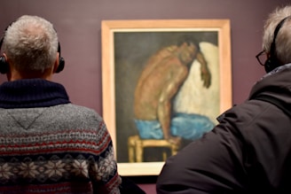 Two individuals with headphones are standing in front of a framed painting. The artwork depicts a seated figure with a bare upper body and blue pants, resting its head on an arm against a dull background. The individuals are viewing the painting, suggesting an art exhibit setting.