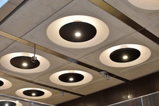 A series of circular recessed ceiling lights with black central panels and bright white illumination are set into a grid-patterned ceiling. Metallic elements and modern design features are highlighted, with variations in light intensity across different fixtures. The surroundings suggest an industrial or contemporary architectural space.