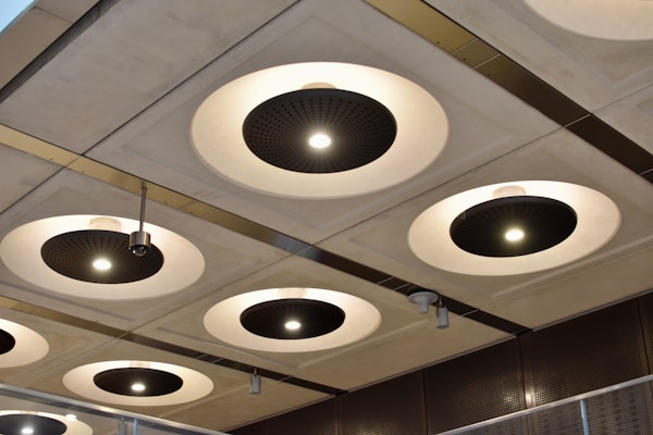 A series of circular recessed ceiling lights with black central panels and bright white illumination are set into a grid-patterned ceiling. Metallic elements and modern design features are highlighted, with variations in light intensity across different fixtures. The surroundings suggest an industrial or contemporary architectural space.