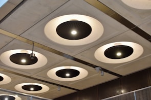 A series of circular recessed ceiling lights with black central panels and bright white illumination are set into a grid-patterned ceiling. Metallic elements and modern design features are highlighted, with variations in light intensity across different fixtures. The surroundings suggest an industrial or contemporary architectural space.