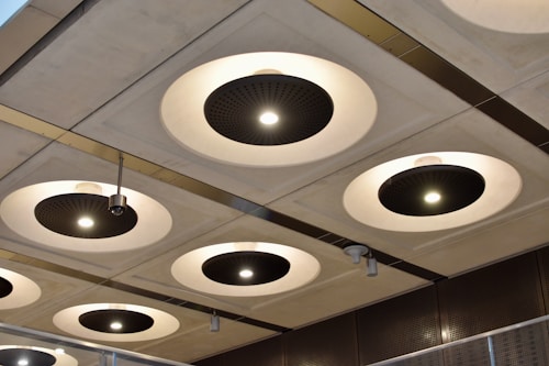 A series of circular recessed ceiling lights with black central panels and bright white illumination are set into a grid-patterned ceiling. Metallic elements and modern design features are highlighted, with variations in light intensity across different fixtures. The surroundings suggest an industrial or contemporary architectural space.