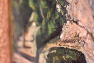 A close-up of an artwork featuring abstract forms with green and brown hues. The brush strokes create a textured surface evoking natural elements like foliage and rocks.