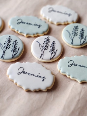 Several decorated cookies laid out on a beige surface. Some cookies have a wavy edge and contain a handwritten name, 'Jeremias,' while others have a smooth edge featuring a simple black line drawing of a plant with leaves.