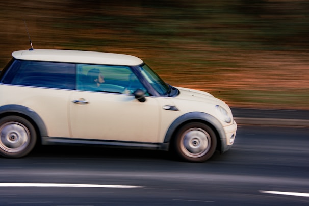 A cream-colored compact car is moving quickly along a roadway with blurred background indicating motion. The driver is visible inside the vehicle, which features a distinctive rounded shape.