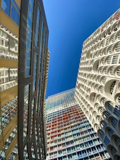 Several modern high-rise buildings with unique architectural designs are seen from a low angle against a clear blue sky. The building on the left has a reflective surface with glass windows, while the building on the right features white arches. The central building showcases a grid pattern with multicolored panels.
