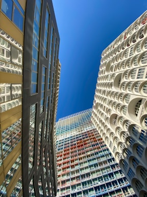 Several modern high-rise buildings with unique architectural designs are seen from a low angle against a clear blue sky. The building on the left has a reflective surface with glass windows, while the building on the right features white arches. The central building showcases a grid pattern with multicolored panels.