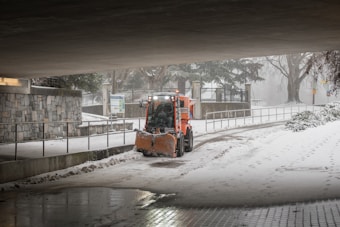 A snow plow clears snow from a pathway beneath an overpass amid a snowy landscape. The plow is bright orange and contrasts with the muted gray and white surroundings. Snow continues to fall, creating a tranquil and wintry scene. The surrounding area includes a stone wall, leafless trees, and some signage in the background.