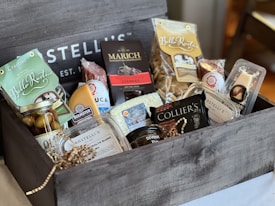 A wooden gift box filled with assorted gourmet food items including cheese, crackers, dried fruits, chocolates, and preserves. The box features brands like Bello Rustico, Marich, and Beemster. The items are attractively arranged with some packaging in clear plastic, others in paper, and the box is accented with decorative straw.