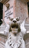 A stone sculpture of a fierce, winged lion with its mouth open, displaying its teeth prominently. The statue is weathered, with visible signs of aging and patina. Behind the sculpture, there is a stone monument with an inscription displaying text in Portuguese.