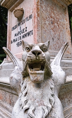 A stone sculpture of a fierce, winged lion with its mouth open, displaying its teeth prominently. The statue is weathered, with visible signs of aging and patina. Behind the sculpture, there is a stone monument with an inscription displaying text in Portuguese.