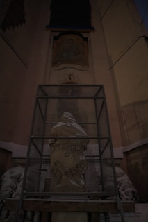 A statue is covered with protective material and enclosed in a metal cage, set against an architectural backdrop with ornate details and inscriptions.