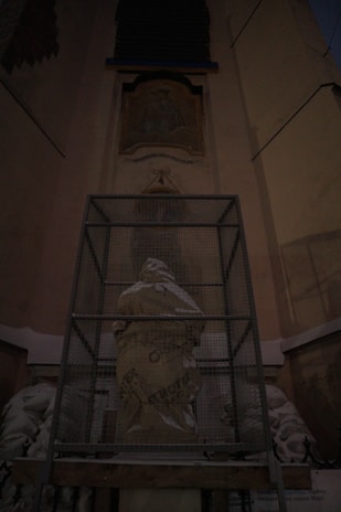 A statue is covered with protective material and enclosed in a metal cage, set against an architectural backdrop with ornate details and inscriptions.