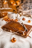 A warmly lit scene featuring a decorative Holy Bible with intricate designs on the cover. The Bible is resting on a soft textured surface, surrounded by small orange Christmas ornaments and fairy lights. A pen is placed on top of the Bible. In the background, a ceramic mug with a holiday design and a cozy cushion are visible, contributing to a festive and cozy atmosphere.