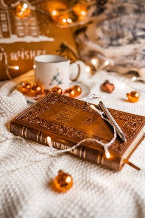 A warmly lit scene featuring a decorative Holy Bible with intricate designs on the cover. The Bible is resting on a soft textured surface, surrounded by small orange Christmas ornaments and fairy lights. A pen is placed on top of the Bible. In the background, a ceramic mug with a holiday design and a cozy cushion are visible, contributing to a festive and cozy atmosphere.