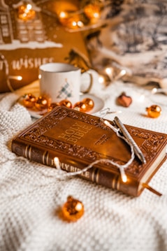 A warmly lit scene featuring a decorative Holy Bible with intricate designs on the cover. The Bible is resting on a soft textured surface, surrounded by small orange Christmas ornaments and fairy lights. A pen is placed on top of the Bible. In the background, a ceramic mug with a holiday design and a cozy cushion are visible, contributing to a festive and cozy atmosphere.