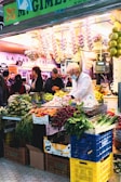 A bustling market scene with a vendor wearing a white coat and mask tending to a vibrant display of fresh produce. The stand is filled with a variety of vegetables including carrots, radishes, and greens. Bunches of garlic and other items hang above the stall. Several people are visible in the background, contributing to a lively atmosphere.