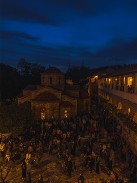 A nighttime scene of a gathering in front of an illuminated, historic stone church with a domed roof. Numerous people stand and casually interact in the courtyard, which is surrounded by trees and a building with archways glowing with warm light.