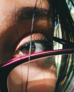 A close-up view of an eye with long eyelashes, partially obscured by strands of black hair. The eye is framed by the rim of a purple or dark pink eyeglass frame. Soft natural light casts shadows across the skin, highlighting the textures and details.