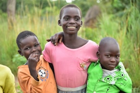 Three children are standing closely together outdoors, each with an arm around the other, conveying a sense of camaraderie and friendship. The child in the center is wearing a pink shirt and smiling broadly, while the children on either side wear orange and green shirts, also smiling softly. The background consists of tall green grass and trees, suggesting a rural or natural setting.