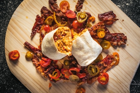 A neatly arranged burrito cut in half with a filling of scrambled eggs and other ingredients is placed on a wooden board. Surrounding the burrito are crispy strips of bacon and slices of colorful bell peppers and jalapeños, adding a pop of color to the presentation.
