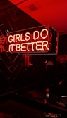 A neon sign with pink and red colors illuminating the words 'Girls Do It Better' against a dark tiled background. The glow of the neon lights casts a warm, vibrant hue on the surrounding surfaces.